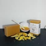 Savon Rayon de Miel Milléniale avec étiquette et sans étiquette, accompagné de copeaux de cire d’abeille et d’une soucoupe de miel, sur ardoise noire.