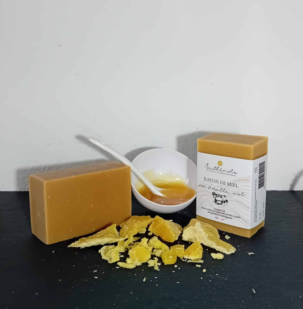 Savon Rayon de Miel Milléniale avec étiquette et sans étiquette, accompagné de copeaux de cire d’abeille et d’une soucoupe de miel, sur ardoise noire.